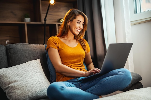 Inquadratura di una giovane donna attraente seduta a gambe incrociate sul divano e che usa il suo laptop a casa.