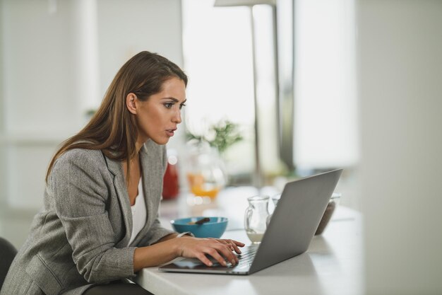 Inquadratura di una giovane donna d'affari che usa il suo laptop mentre fa colazione nella sua cucina durante il lavoro da casa.