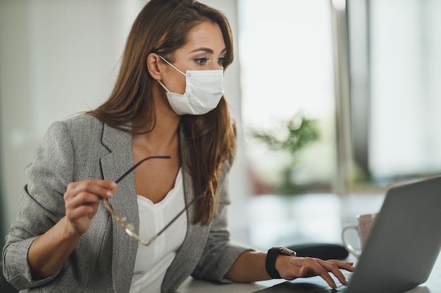 Inquadratura di una giovane donna d'affari con maschera protettiva seduta da sola nel suo ufficio e che lavora su un laptop durante la pandemia del virus corona.