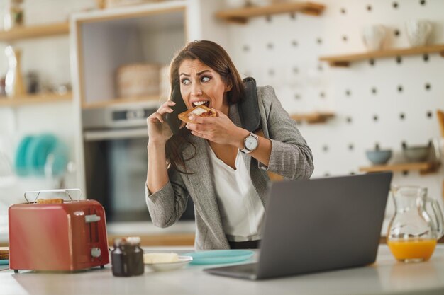 Inquadratura di una giovane donna d'affari multi-tasking che fa colazione e parla sullo smartphone nella sua cucina mentre si prepara per andare al lavoro.