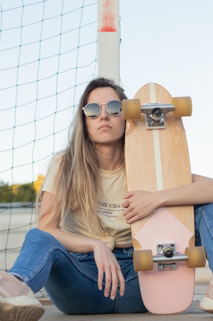 Inquadratura frontale di una ragazza bionda ribelle seduta sul pavimento accanto al suo skateboard vintage rosa