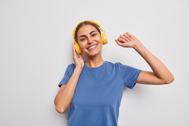 Inquadratura in vita di una donna europea spensierata positiva che balla e cattura ogni brano di musica indossa una maglietta blu casual ascolta la canzone preferita tramite cuffie wireless isolate su sfondo bianco.