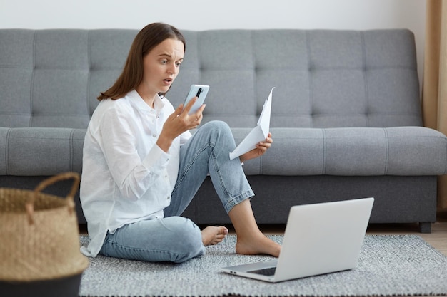 Inquadratura interna di una donna stupita e stupita che indossa una camicia bianca e jeans seduta sul pavimento e guardando lo schermo del cellulare con stupore tenendo la carta in mano in posa vicino al laptop con divano sullo sfondo