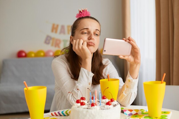 Inquadratura interna di una donna triste sconvolta che indossa abiti bianchi seduta a tavola con torta e bevanda, tiene in mano il telefono cellulare, guarda video, prova dolore per festeggiare da sola.