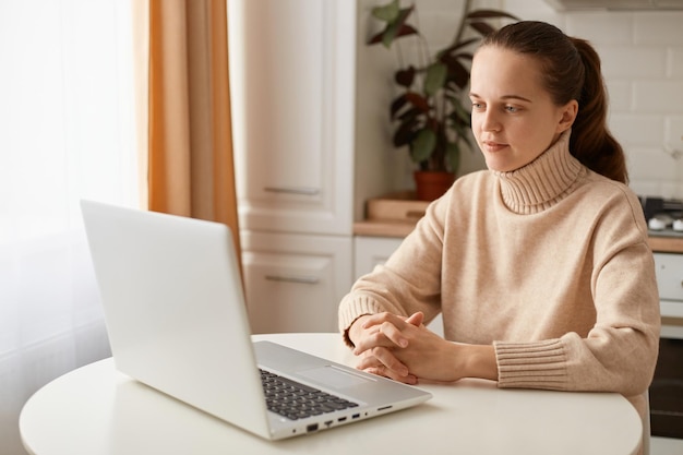 Inquadratura interna di una giovane donna attraente e calma che indossa un maglione beige seduto in cucina con un notebook che lavora tenendo una lezione online guardando il display del laptop con un'espressione seria