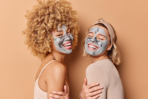 Inquadratura laterale di donne felici vicine l'una all'altra hanno espressioni allegre applicare maschere di bellezza per il trattamento della pelle sorriso ampiamente isolato su sfondo marrone Benessere e cura del viso