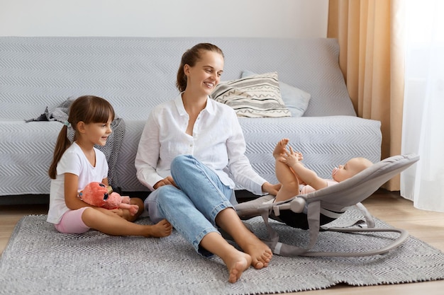 Inquadratura orizzontale di adorabile donna felice che indossa una camicia bianca e jeans seduta sul pavimento con i suoi bambini che trascorrono del tempo con un bambino sulla sedia a dondolo insieme al bambino più grande