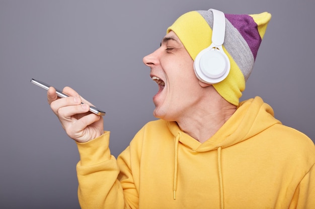 Inquadratura orizzontale di attraente uomo allegro felice in felpa con cappuccio gialla e cuffie wireless che canta canzoni tenendo il messaggio di registrazione dello smartphone con la sua canzone in posa isolato su sfondo grigio