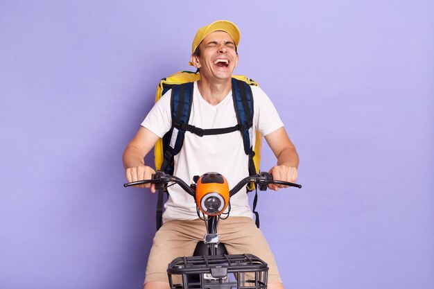 Inquadratura orizzontale di divertente uomo di consegna felice di cibo da asporto su scooter che consegna cibo e ridendo di buon umore godendosi il suo lavoro in posa isolato su sfondo viola