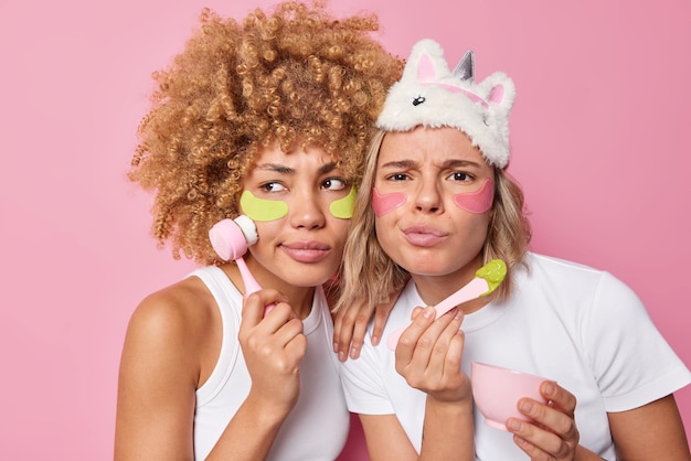 Inquadratura orizzontale di donne serie prendersi cura dell'aspetto applicare patch di bellezza idrogel utilizzare massaggiatore viso per la cura della pelle applicare maschera facciale stand uno accanto all'altro isolato su sfondo rosa