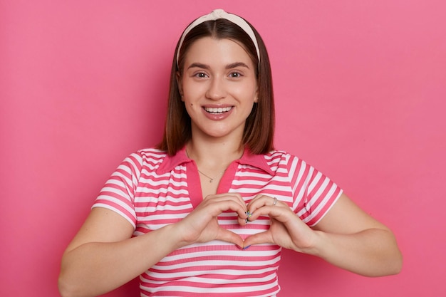 Inquadratura orizzontale di felice romantica giovane donna che indossa maglietta a righe e fascia per capelli che mostra il gesto del cuore con le mani che esprimono amore e posa gentile isolata su sfondo rosa