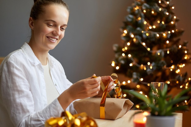 Inquadratura orizzontale di sorridente adorabile femmina imballaggio regalo di Natale con fiocco d'oro sullo sfondo dell'albero di Natale con luce avvolgente regali per la sua famiglia e gli amici per le vacanze di Capodanno