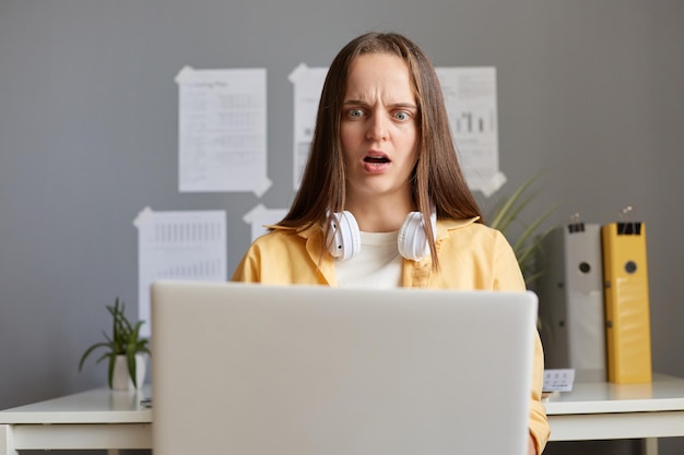 Inquadratura orizzontale di una donna caucasica spaventata e scioccata con i capelli castani che lavora al computer portatile in ufficio seduto e guardando lo schermo del notebook con grandi occhi che hanno problemi con il lavoro o lo studio