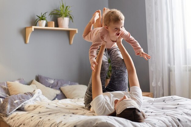 Inquadratura orizzontale di una donna con la sua bambina che gioca in camera da letto mentre giace sul letto madre che lancia il suo bambino in aria divertendosi insieme al mattino