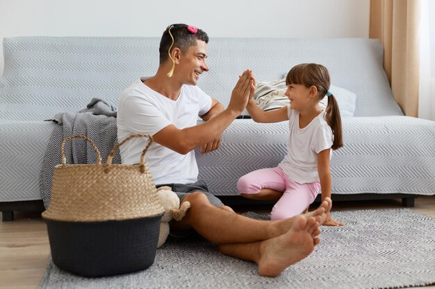 Inquadratura orizzontale di uomo divertente con accessori per bambini sui capelli padre che indossa una maglietta bianca in stile casual e jeans short seduto sul pavimento con sua figlia che si dà cinque a vicenda