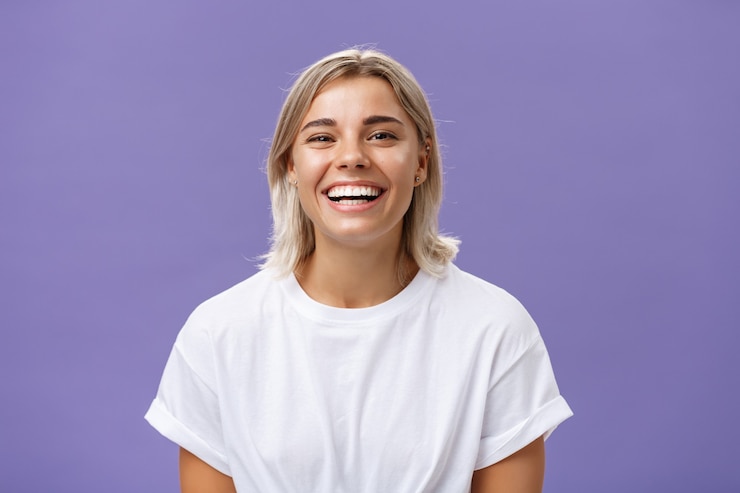 Foto inquadratura ravvicinata di gioiosa e affascinante femmina bionda con un sorriso lieto e compiaciuto in piedi in t-shirt bianca, trascorrere del tempo in una fantastica compagnia divertente
