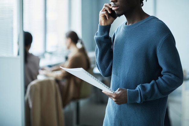 Inquadratura ritagliata in blu di un giovane afroamericano che parla al telefono in ufficio e tiene in mano d