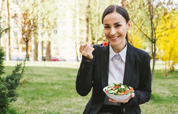 Insalata al pranzo di lavoro Ritratto di una bella donna d'affari moderna e sorridente in un abito nero con un piatto di insalata di verdure nelle mani che mangia per strada vicino al lavoro