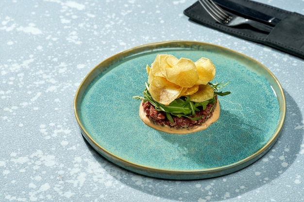 Insalata appetitosa - tartare di vitello con salsa, rucola, patatine in un piatto blu su sfondo blu