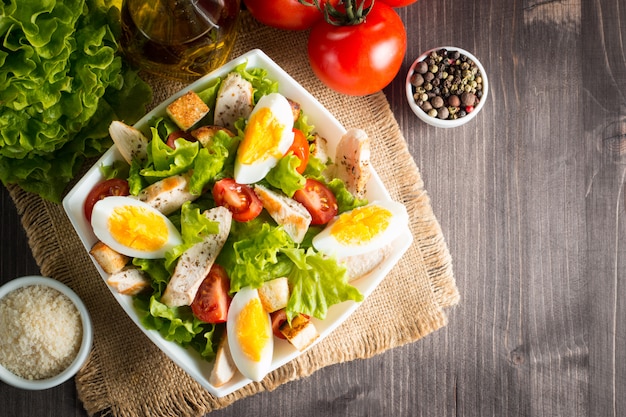 Insalata Caesar fresca con delizioso petto di pollo, rucola, spinaci, cavolo, rucola, uovo, parmigiano e pomodoro ciliegia su fondo di legno. Olio, sale e pepe Concetto di cibo sano e dieta.