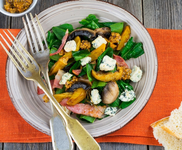 Insalata calda autunnale con patate, funghi, pancetta, spinaci, salsa di senape e formaggio su tovagliolo arancione. Fondo in legno vecchio. Messa a fuoco selettiva.