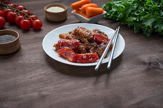 Insalata calda di melanzane e pomodori in stile coreano con semi di sesamo ed erbe. Cibo asiatico. Piatto vegetariano. Sfondo di legno