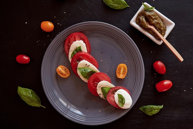 Insalata caprese mediterranea italiana con mozzarella pomodori foglie di basilico e salsa di pesto su fondo rustico nero Cibo piatto