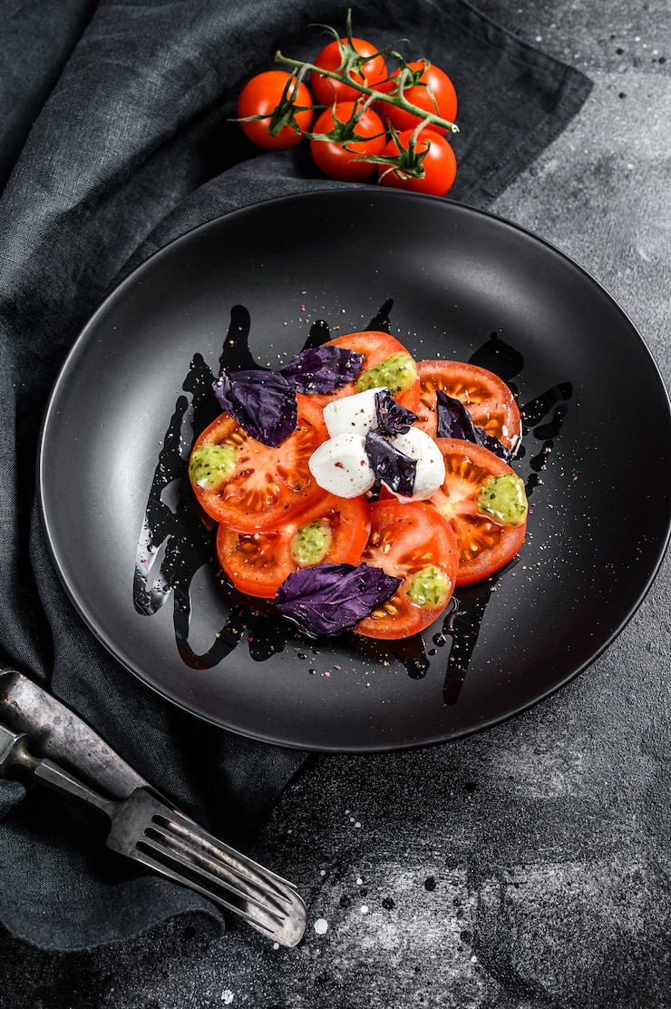 Foto insalata caprese tradizionale italiana. pomodorini, mozzarella, basilico, pesto. sfondo nero. vista dall'alto