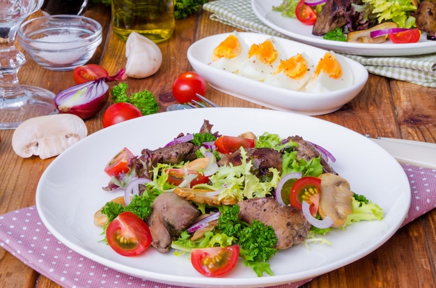 Insalata con fegato di pollo, funghi fritti e pomodori su un legno scuro.