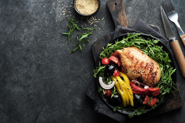 Insalata con filetto di bres di pollo alla griglia con rucola fresca pomodori e olive su piatto nero su vecchio sfondo scuro Carne di pollo con insalata Pranzo chetogenico Cibo sano Vista dall'alto