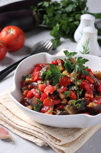 Insalata con melanzane, pomodori, aglio e prezzemolo in una ciotola bianca su sfondo grigio chiaro. Formato verticale