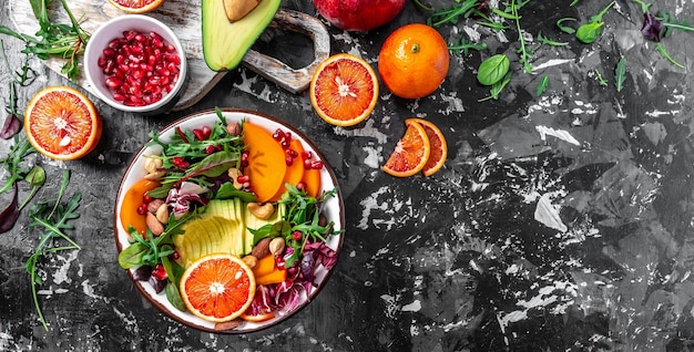 Insalata di buddha vegetariano sano con avocado cachi arancia rossa noci spinaci rucola e melograno su uno sfondo scuro Formato banner lungo vista dall'alto