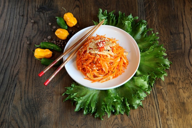 Insalata di carne di calamaro coreano di carota in un piatto con bacchette su tavola di legno di quercia e foglie di insalata verde, pepe giallo fresco. Insalata di carote piccante in stile coreano su uno sfondo scuro. Cucina Koryo-saram.