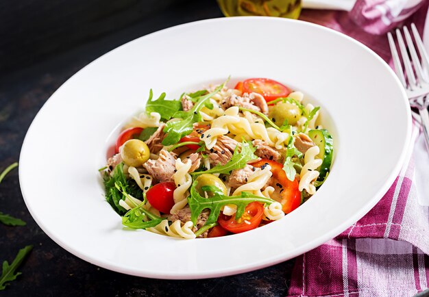 Insalata di pasta con tonno, pomodori, olive, cetriolo, peperone dolce e rucola su fondo rustico.