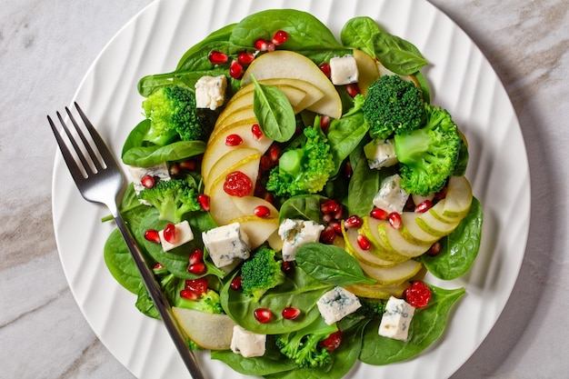 Insalata di pere con cubetti di formaggio blu, broccoli, spinaci, mirtilli rossi secchi e semi di melograno su un piatto su un tavolo di marmo, contorno del ringraziamento, vista orizzontale dall'alto, primo piano