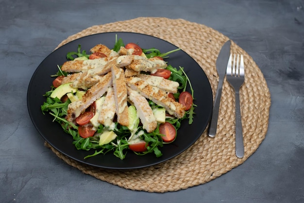 Insalata di pollo con avocado, rucola e pomodorini in un piatto nero su sfondo grigio pietra.