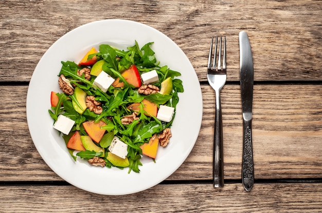 Insalata di rucola, cachi, noci, avocado, formaggio feta su un piatto bianco su un tavolo di legno