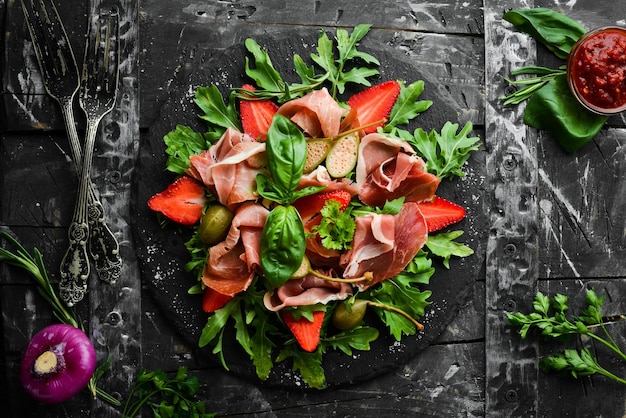 Insalata di rucola prosciutto fragole e capperi su una lastra di pietra nera Cucina italiana