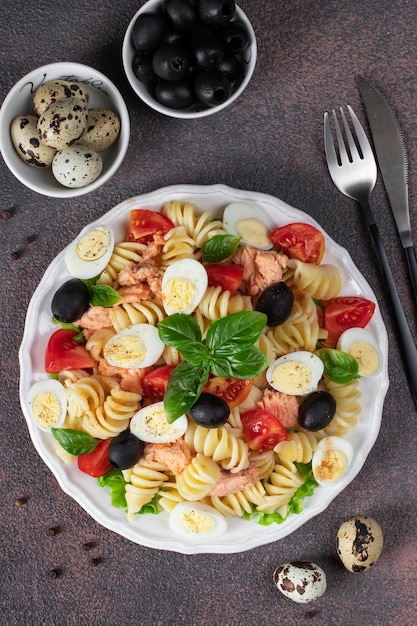 Insalata di salmone in scatola con pasta pomodorini uova di quaglia basilico e olive nere su fondo marrone