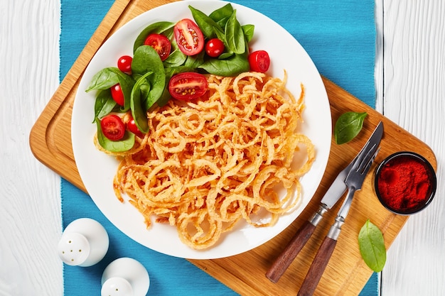 Insalata di spinaci freschi e pomodori serviti con anelli di cipolla fritti croccanti francesi e stringhe su un piatto bianco su una tavola di legno, vista dall'alto, piatto