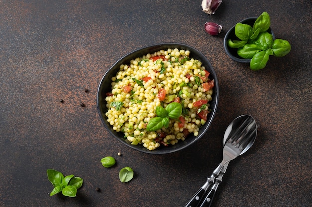 Insalata di verdure con pasta ptim o Birdy cuscus israeliano decorato con foglie di basilico su sfondo marrone scuro Vista dall'alto