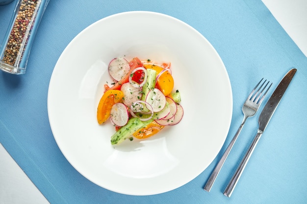 Insalata di verdure fresca e leggera di pomodori, cetrioli e ravanelli in un piatto bianco sulla tovaglia blu.