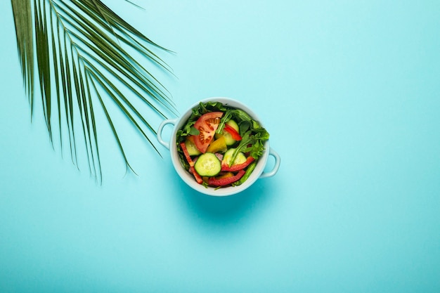 insalata di verdure in una ciotola blu e una foglia verde di una palma su una superficie blu.