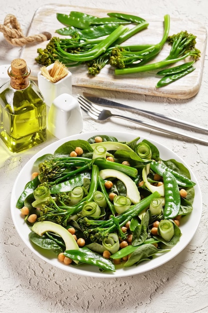 Insalata di verdure verdi di broccoli saltati piselli avocado cetriolo ceci spinaci con condimento all'olio d'oliva servito su un piatto bianco con posate su un tavolo bianco vista dall'alto