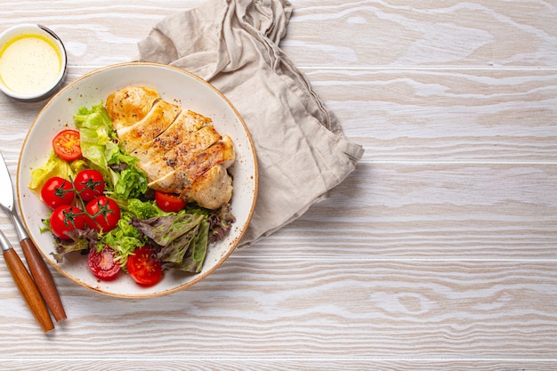 Insalata di verdure verdi sane con filetto di petto di pollo alla griglia su piatto di ceramica con olio d'oliva sul lato sul tavolo da cucina in legno bianco vista dall'alto piatto disteso, concetto di cibo dietetico con spazio per il testo