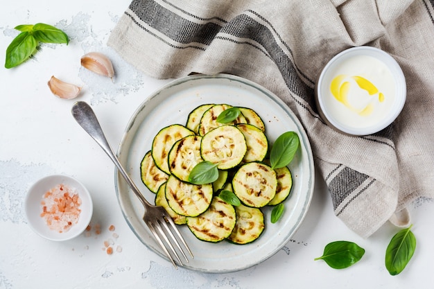 Insalata di zucchine grigliate con foglie di basilico, salsa allo yogurt e pane fritto in un semplice piatto di ceramica su una superficie di cemento bianco