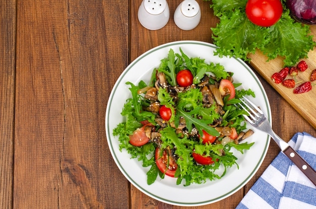 Insalata dietetica di foglie di rucola, pomodori e funghi fritti su un tavolo di legno.