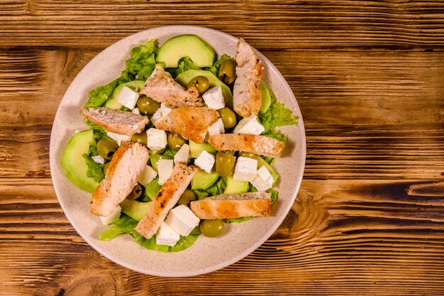 Insalata fresca con carne di pollo, formaggio feta, avocado, olive verdi e foglie di lattuga in piatto di ceramica su tavola in legno rustico. Vista dall'alto