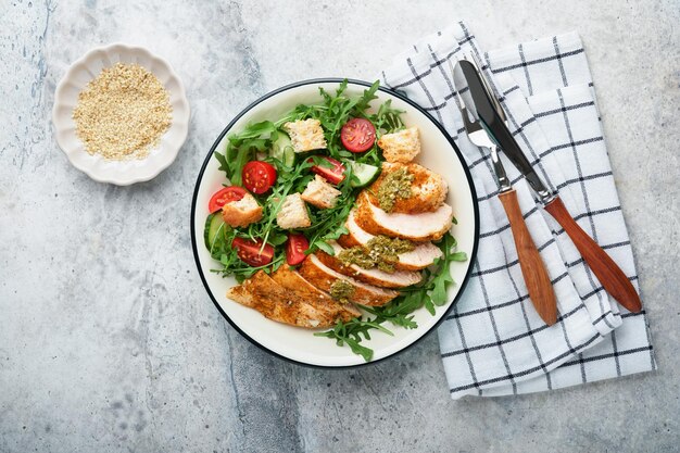 Insalata fresca con filetto di pollo alla griglia, rucola, pomodori, cetriolo e salsa al pesto, semi di sesamo con olio d'oliva in una ciotola bianca su sfondo ardesia chiaro, menu pranzo sano, cibo dietetico, vista dall'alto