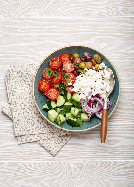 Insalata greca mediterranea con pomodori, formaggio feta, cetriolo, olive intere e cipolla rossa in piatto di ceramica blu su fondo di legno bianco dall'alto, antipasto tradizionale della Grecia